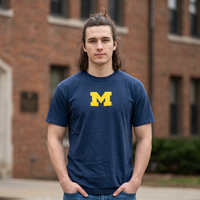 Person wearing a navy blue t-shirt with a yellow 'M' logo in front of a brick building.
