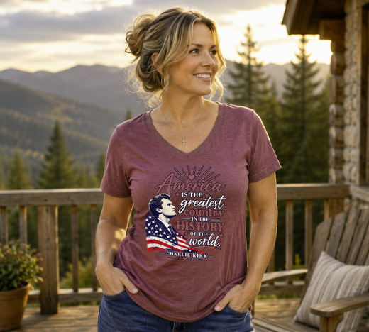 A woman stands on a wooden porch wearing a heathered mauve V-neck graphic t-shirt with a patriotic design featuring a silhouette of a woman and the text 'America is the greatest country in the history of the world.' by Charlie Kirk.